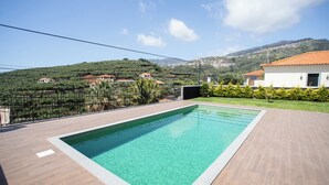 A swimming pool with a wooden deck, surrounded by a fence, and a view of a hillside with houses. - Dori’s Modern Home w/Pool & Sea & Mountain Views — 15 Min to City Center (Funchal)