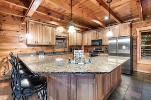 A kitchen with wooden cabinets, granite countertops, a stainless steel refrigerator, and a bar area with stools. - Timber Sky Lodge! Elevator/Pool/Theater | Sleep 27 (Sevierville)