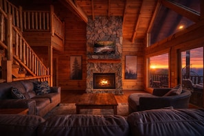 A log cabin interior with a stone fireplace, leather furniture, a staircase, and a view of a sunset. - Timber Sky Lodge! Elevator/Pool/Theater | Sleep 27 (Sevierville)