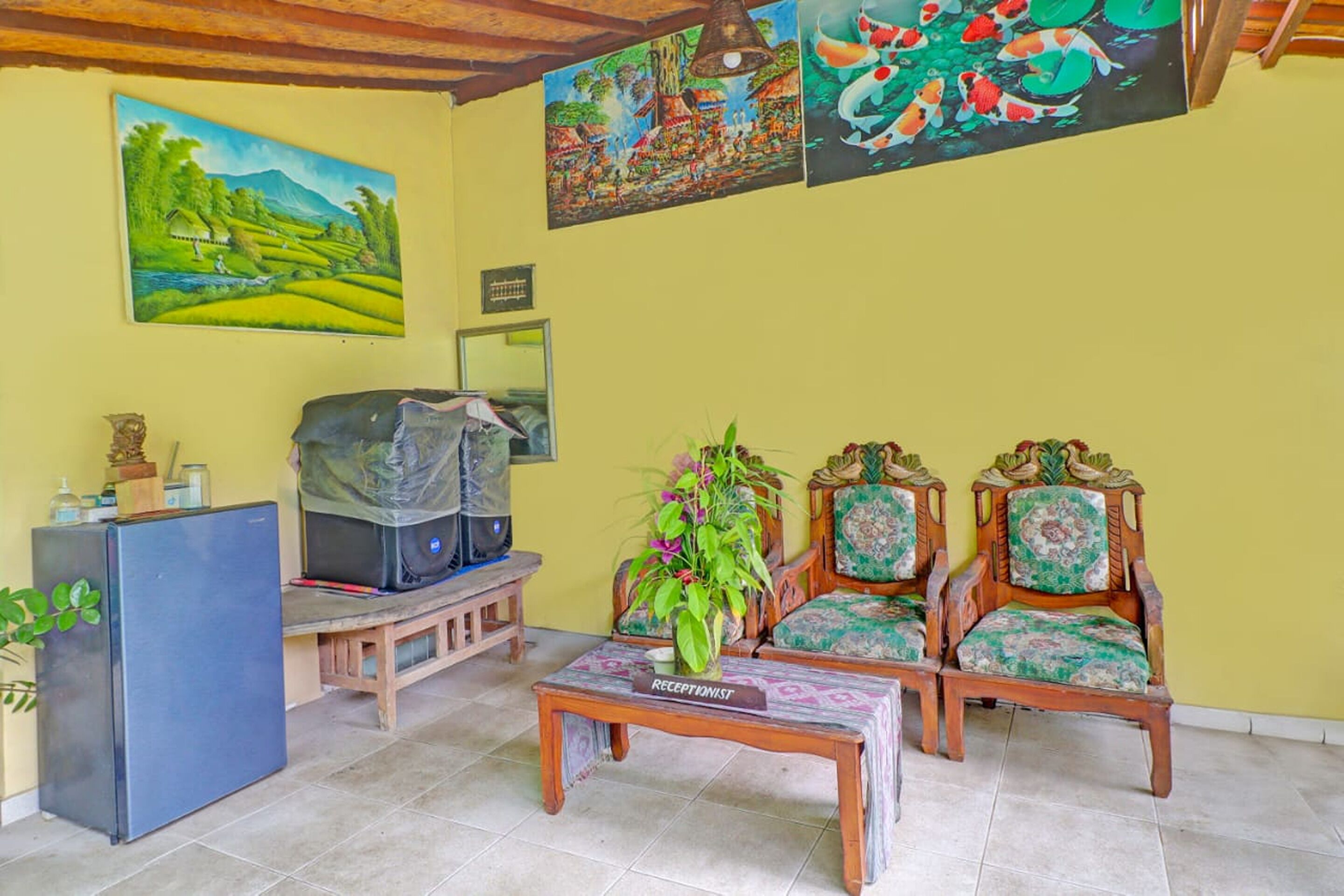 A living room with a reception desk, two wooden chairs, a coffee table, and a refrigerator.