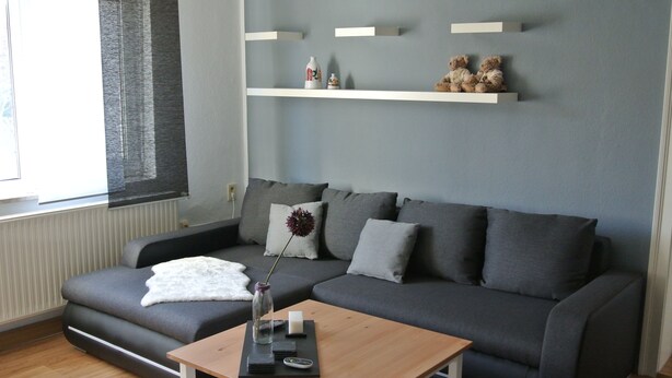 Een moderne woonkamer met een grijze bank, een houten salontafel en een wit vloerkleed. Boven de bank hangen twee planken met decoratieve objecten.