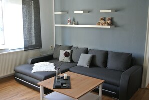 Ett modernt vardagsrum med en grå soffa, ett soffbord i trä och en vit matta. Ovanför soffan finns två hyllor med dekorativa föremål.