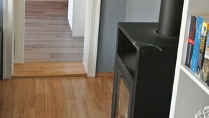 Een kamer met een houtkachel, een boekenplank vol boeken en uitzicht op een keuken met rode kasten en een wit aanrecht.