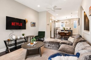 A modern living room with a flat-screen TV, a sofa, a coffee table, and a dining area in the background. - Premium house for large groups with sky deck view! (Nashville)