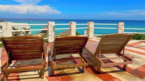 Three wooden lounge chairs facing a coastal view with a white railing and a stone pillar. - Cozy Studio apartment in tranquil Cancún with AC (Cancún)