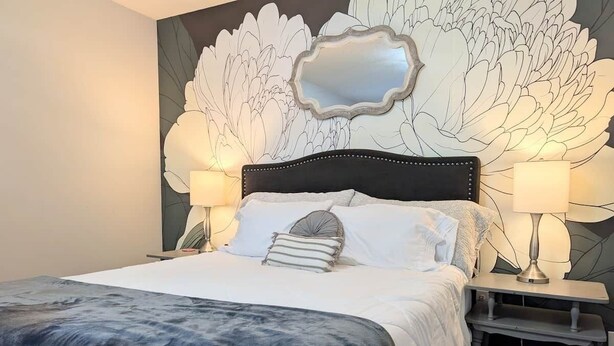 A neatly made bed with a dark blanket, two bedside lamps, and a wall with a large floral design. - Southern Magnolia 3BR/2Bath Home 5 min to base (Enterprise)
