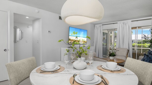 A modern dining room with a round marble table set for four, white chairs, and a large window with a view of a beach. - Beach Luxury Retreat • Walk to Beach & 5th Ave (Naples)