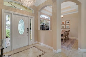 Ein geräumiges Foyer mit einer großen weißen Tür, einem Essbereich mit Stühlen und einer Chandelier.
