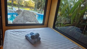 A room with a bed, a window with a view of a swimming pool and rocks, and a window blind. - Cabanas.tur Ilhabela (Ilhabela)