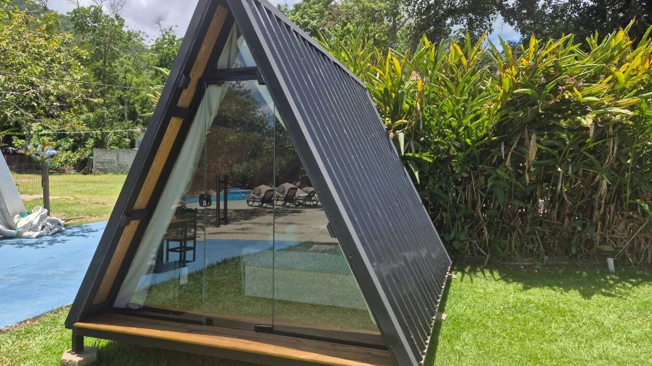 Una estructura de vidrio triangular con techo de metal, rodeada por un área verde y follaje denso.