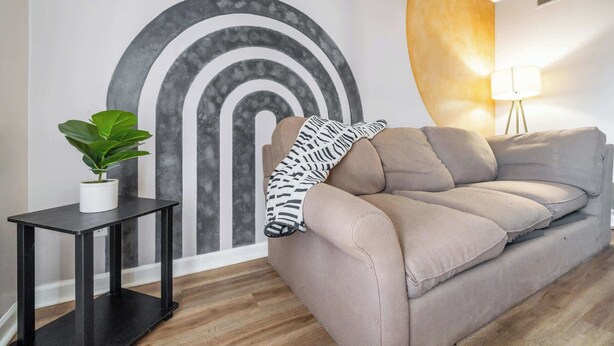 A living room with a beige sofa, a black side table with a potted plant, a wall with a large abstract mural, and a floor lamp.