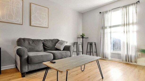 A modern living room with a grey sofa, a wooden coffee table, two bar stools, and a large window with sheer curtains. - Spacious Town Home | Free Parking | Pet Friendly (Louisville)