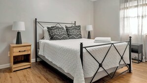 A neatly made bed with a white quilt, two patterned pillows, and a matching throw. There is a wooden nightstand with a lamp, a small drawer, and a bedside table with a lamp. The room has a wooden floor and a window with sheer curtains. - Spacious Town Home | Free Parking | Pet Friendly (Louisville)