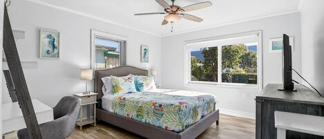 A bedroom with a bed, a desk, a chair, a ceiling fan, and a window with a view of greenery.