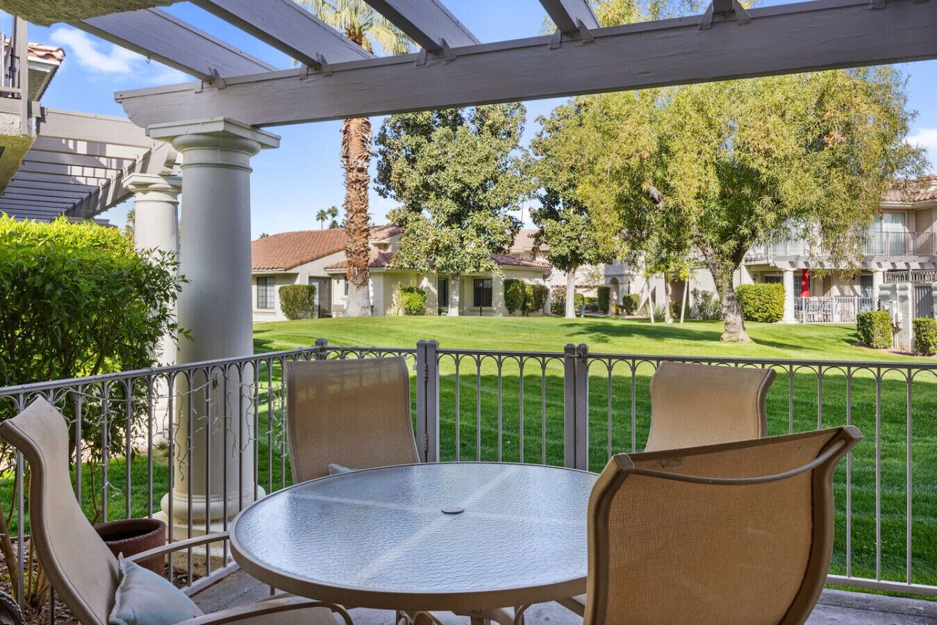 A covered patio with a round table and chairs, a view of a well-maintained lawn and houses in the background.
