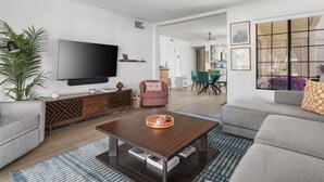 A modern living room with a flat-screen TV, grey sofas, a coffee table, and a dining area with chairs and a table.