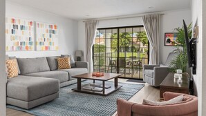 A modern living room with a grey sofa, a coffee table, and a pink armchair. There is a large window with curtains, a painting on the wall, and a plant on the right.