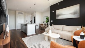 A modern living room with a sofa, coffee table, and a TV. There is a kitchen area with a dining table and chairs. - 2BR Oasis w/ Gym, Parking Near Rvrwalk &Dome (San Antonio)