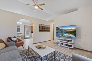 A modern living room with a flat-screen TV, a ceiling fan, and a dining area with a view of the kitchen. - Cute Boise Bench Depot House (Boise)