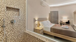 Una habitación de hotel moderna con un espejo grande, una cama con ropa blanca, una mesita de noche de madera y una pequeña mesa con una botella.