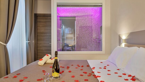 Una habitación de hotel con una cama, una botella de vino y pétalos de rosa sobre la cama.