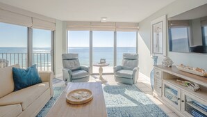 Een ruime woonkamer met een groot raam met uitzicht op zee, een comfortabele bank, twee fauteuils, een salontafel en een tv op een houten kast.