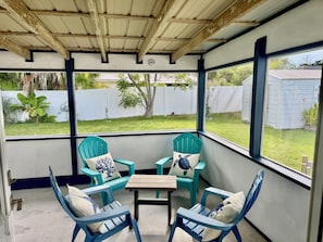 A covered patio with two turquoise chairs, a wooden table, and a view of a backyard. - Ocean Breeze Retreat – Walk to the Beach! (St. Augustine)