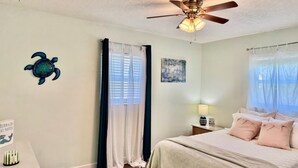 Quarto com cama, ventilador de teto, janela com cortinas, uma obra de arte na parede e uma mesa pequena com abajur.
