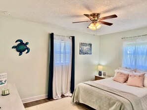A bedroom with a bed, a ceiling fan, a window with curtains, a wall art piece, and a small table with a lamp. - Ocean Breeze Retreat – Walk to the Beach! (St. Augustine)