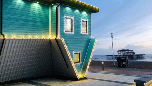 Un edificio color acquamarina con tetto spiovente, illuminato da luci a catena, vicino a una spiaggia con un molo sullo sfondo.