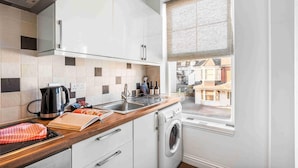 A compact kitchen with a washing machine, a kettle, and a window with a view of buildings. - Queens Cove With Seaviews By My Getaways (Worthing)