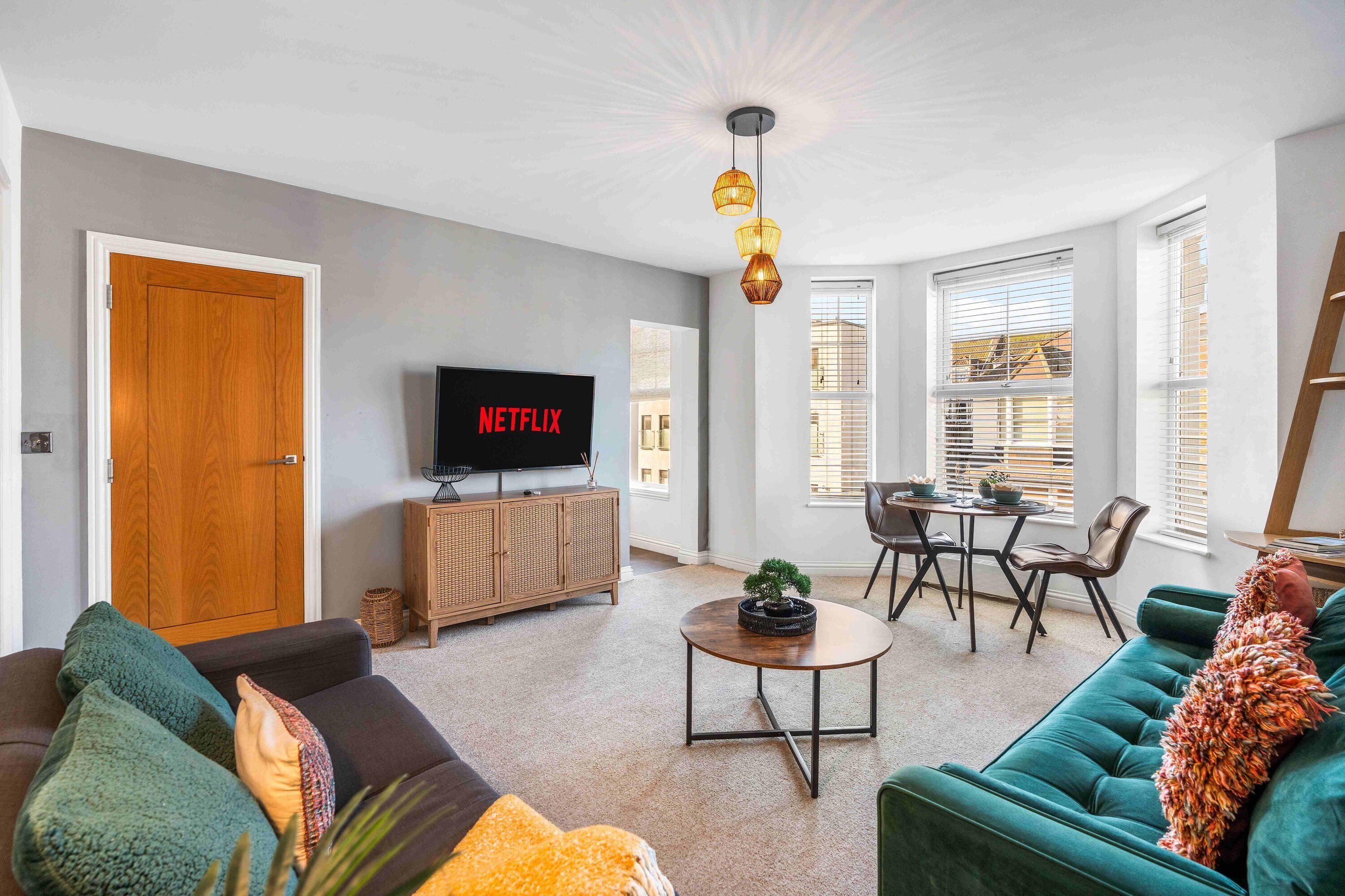 Una sala de estar moderna con un sofá, una mesa de centro y un televisor mostrando 'NETFLIX'. Hay un comedor con una mesa y sillas, y un armario de madera.