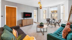 A modern living room with a sofa, a coffee table, and a TV displaying 'NETFLIX'. There is a dining area with a table and chairs, and a wooden cabinet. - Queens Cove With Seaviews By My Getaways (Worthing)