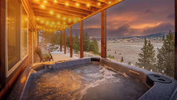 Un jacuzzi bajo una pérgola de madera con luces colgantes, con vistas a un paisaje nevado y un pueblo distante.