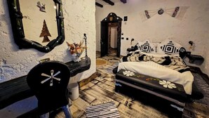 A rustic bedroom with a wooden floor, a bed with patterned bedding, a mirror with a tree design, and a small table with a vase of flowers.