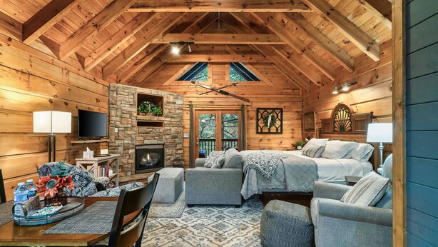 A cozy log cabin interior with a stone fireplace, comfortable seating, and a large window. - Great For Couples.. Great Location! Cozy log cabin w/ NEW Hot Tub! 8 Min to Dtwn (Gatlinburg)