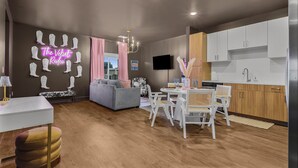 Ein modernes Wohnzimmer mit grauem Sofa, einem Essbereich mit weißen Stühlen, einer Küche mit weißen Schränken und einem Neonlicht an der Wand.