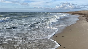 Una playa con olas rompiendo en la orilla, un rompeolas rocoso a lo lejos y un cielo despejado.