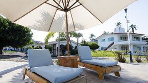 Twee houten lounge fauteuils met kussens onder een grote parasol, geplaatst op een betonnen terras met uitzicht op een wit gebouw en palmbomen in de verte.