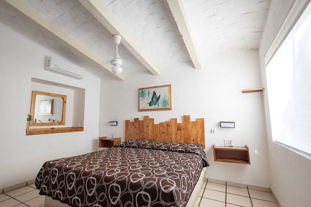 Een slaapkamer met een houten hoofdbord, een bed met een beddegoed met patroon, een plafondventilator, een wandairconditioner, een ingelijste foto en een klein houten plankje.