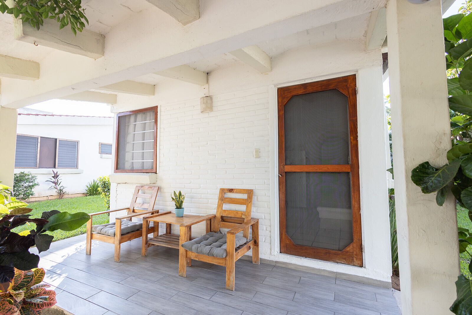 A covered porch with two wooden chairs and a small table, white brick walls, and a large window with a wooden frame.