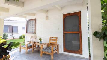 A covered porch with two wooden chairs and a small table, white brick walls, and a large window with a wooden frame.