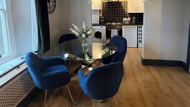 A dining area with a glass table and blue chairs, a kitchen in the background, and a clock on the wall. - garden flat in vibrant area of Clifton  by Bristol university (Bristol)