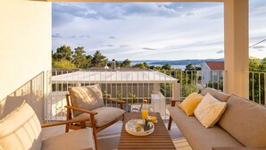 Een balkon met een houten tafel gedekt voor het ontbijt, twee banken en uitzicht op zee en bergen.