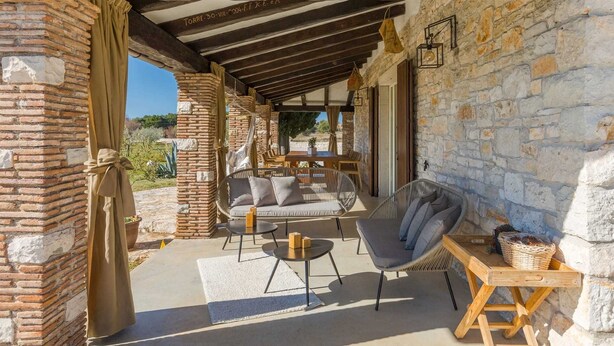 Een terras buiten met stenen muren, een houten pergola, rieten meubels en uitzicht op het landschap.