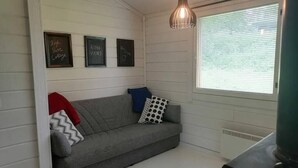 Ein gemütliches Zimmer mit einem grauen Sofa, einer roten Decke und einem schwarzen Heizgerät. An der Wand hängen drei gerahmte Schilder und es gibt ein Fenster mit Jalousien.