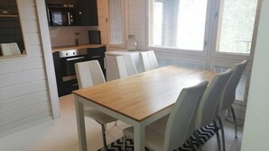 Ein Essbereich mit Holztisch und weißen Stühlen, eine Küche mit modernen Geräten und große Fenster mit Jalousien.