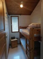 Una habitación pequeña de madera con literas, un pequeño aparador de madera y una ventana con persianas.