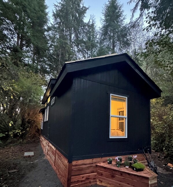 A small, black wooden cabin with a single window, surrounded by dense forest. - The Mossy Hideout Cabin (Klamath)