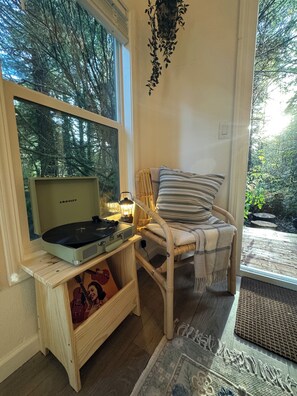 A cozy room with a wooden side table holding a vintage Crosley radio, a wicker chair with a plaid blanket, and a view of a forest through a large window. - The Mossy Hideout Cabin (Klamath)